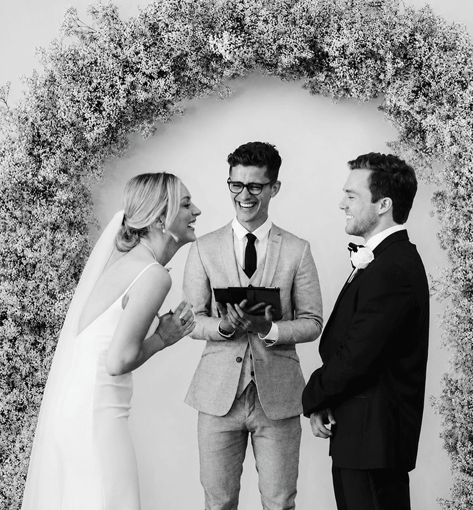 A couple laughing with their officiant beneath a floral arch during the ceremony.