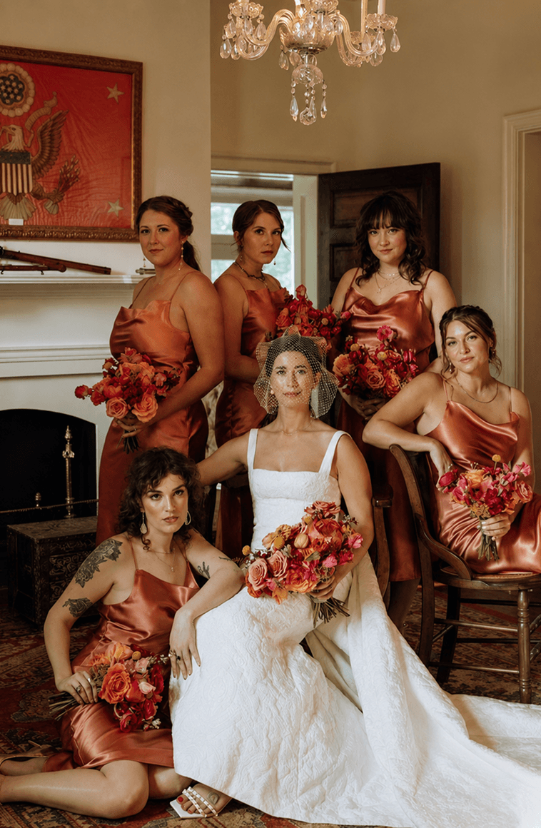 A bride seated among her bridesmaids in rust satin dresses, each holding bouquets.
