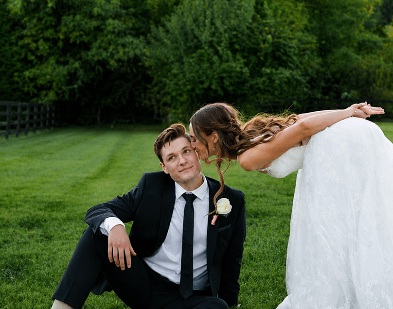 A bride leaning down to kiss her groom as he sits on a lawn.