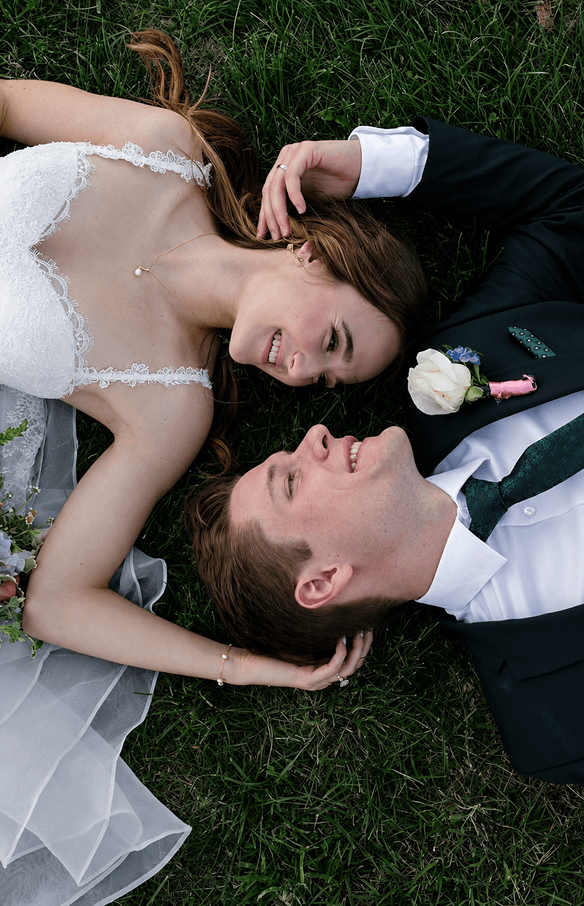A bride and groom lying head to head on the grass, smiling at each other.