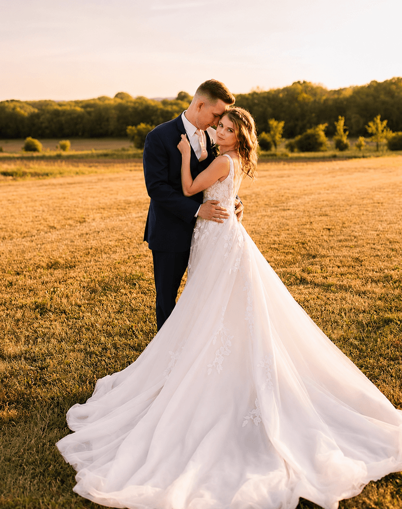 A bride and groom embracing in a golden field at sunset, her train spread on the grass.