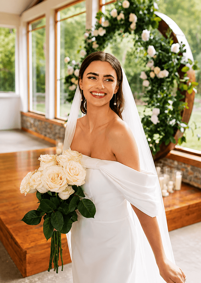 A smiling bride in an off-the-shoulder gown holding a bouquet of white roses.