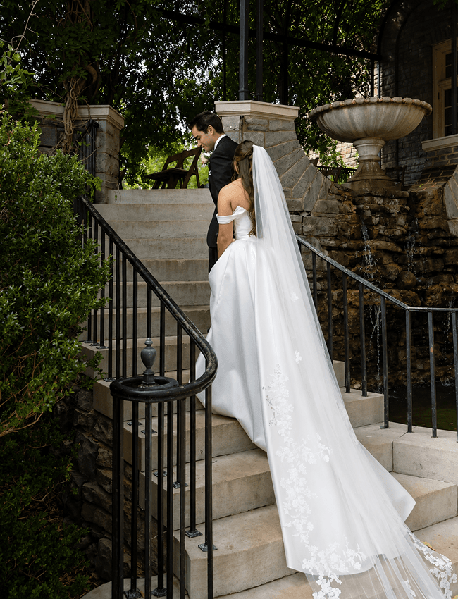 A bride with a long cathedral veil climbing stone steps toward her groom.